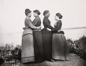 B&W: Two pairs of women in early 1900's dresses embrace lovingly in two pairs.